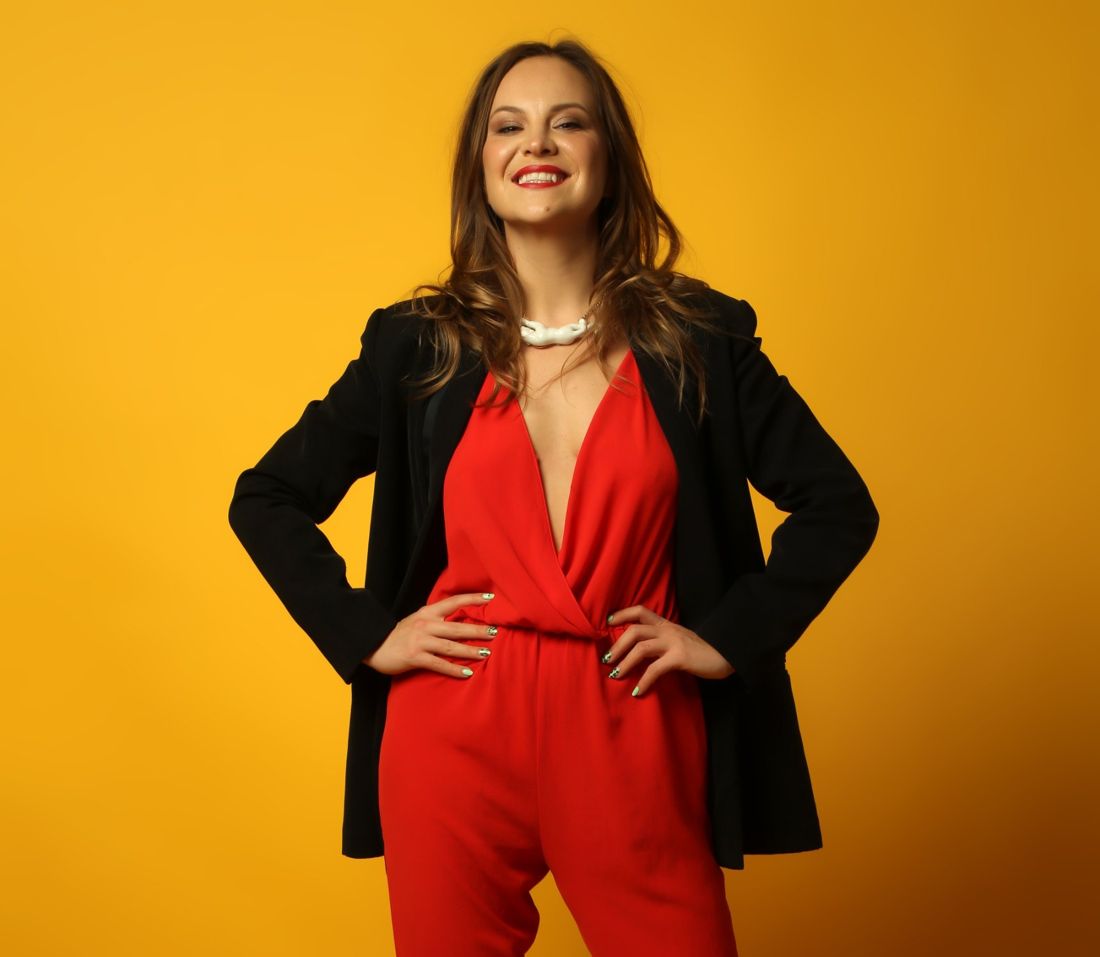 Karina Abramova smiling, hands on hips, in a black blazer against a saffron backdrop