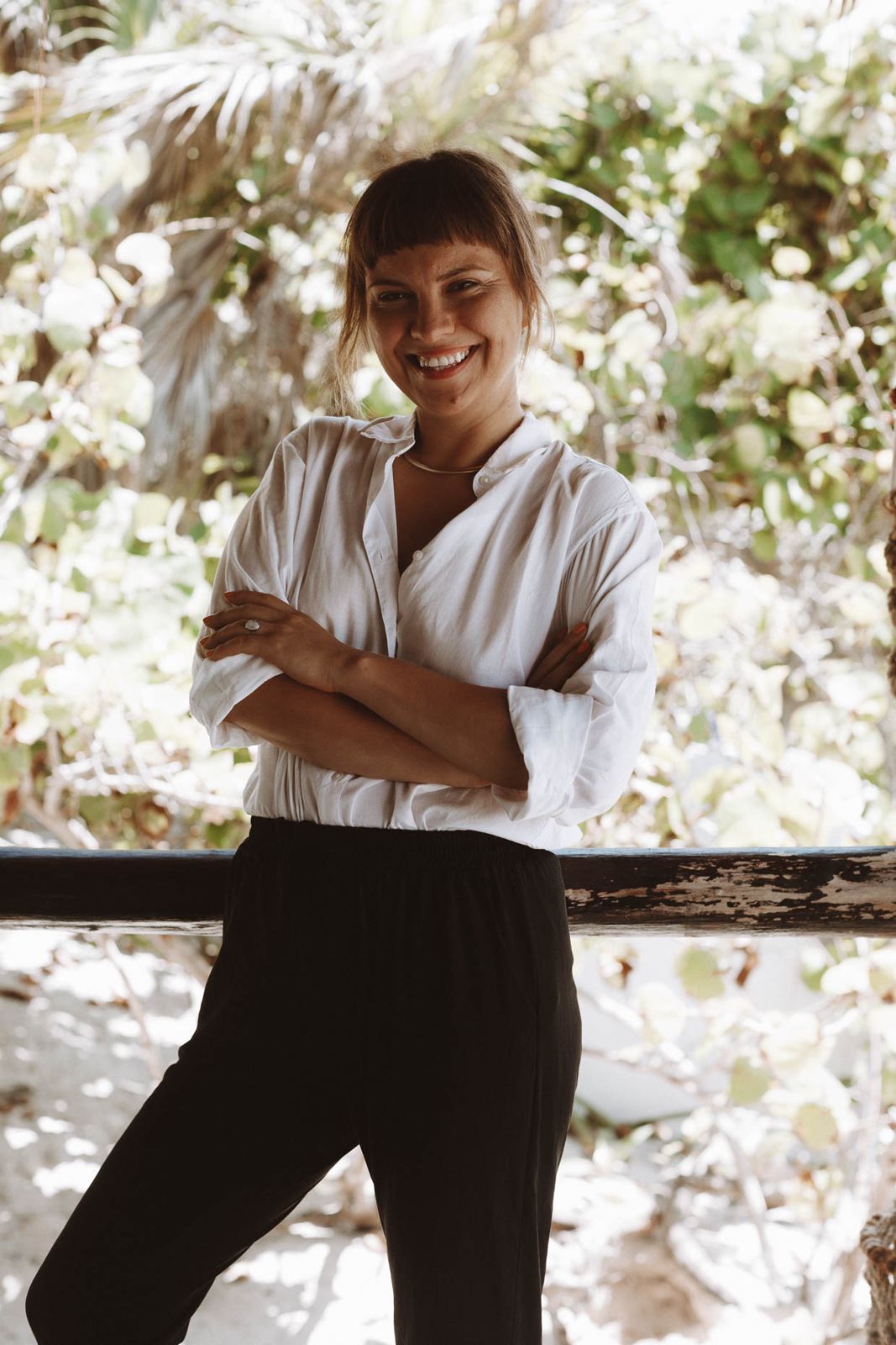 Karina Abramova in a white shirt, arms folded, smiling in front of tropical greenery